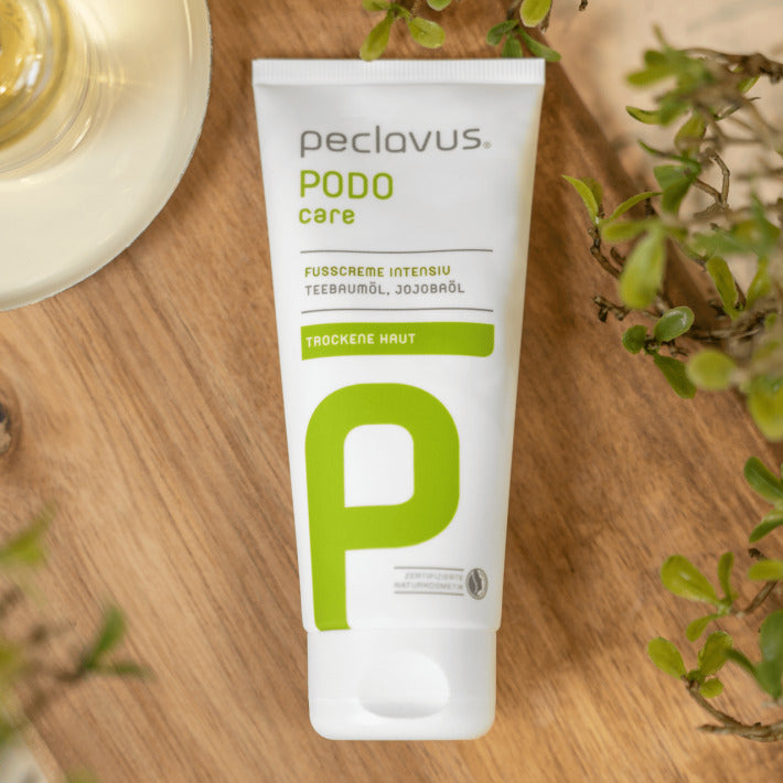 PECLAVUS Fußcreme intensiv – 100ml, Tube auf Holz liegend mit Teebaumblättern