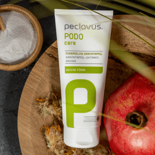 PECLAVUS Fußpeeling Granatapfel – 100ml, Tube liegend auf Holzteller mit Granatapfel und Sandschale