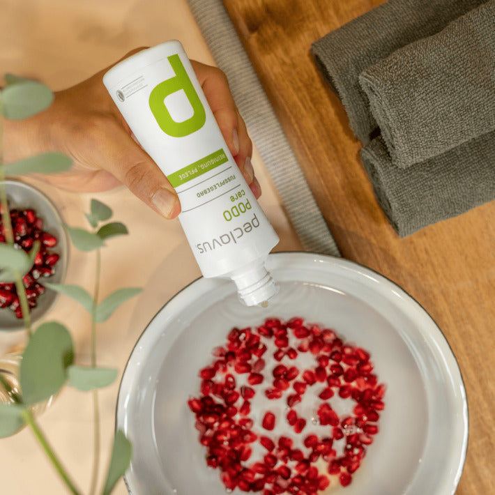 PECLAVUS Fußpflegebad – 150ml, Hand gibt Fußbad in Wasserschale mit Granatapfelkernen