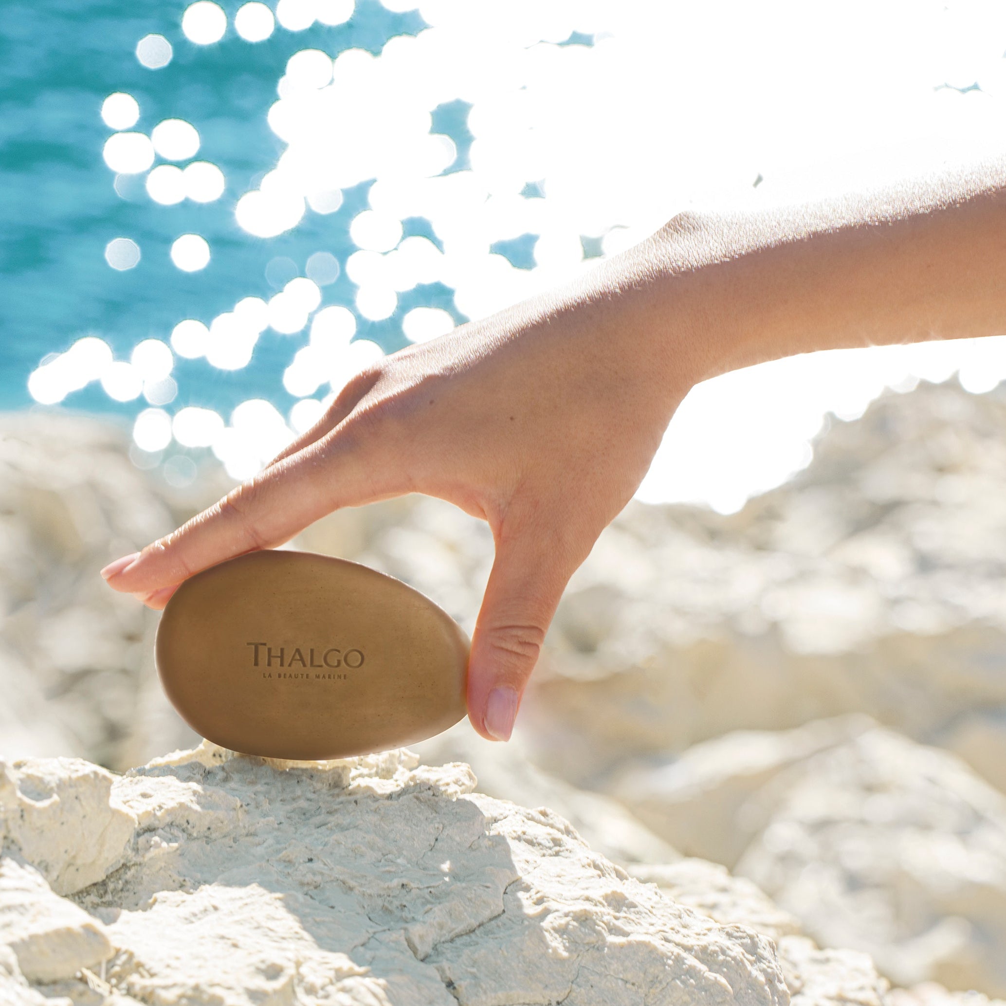 THALGO Éveil à la Mer - Algenseife Gesicht – für alle Hauttypen, 100g, Hand hält Seife vor Steinküste am Meer