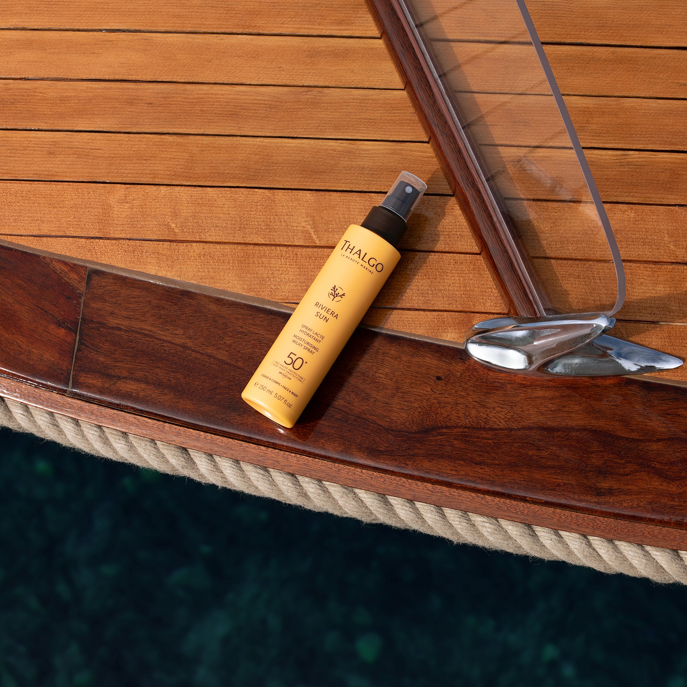 THALGO Riviera Sun Feuchtigkeitsspendendes Sonnenschutz-Spray – LSF 50+, 150ml, Sprühflasche liegend auf Holzboot im Wasser