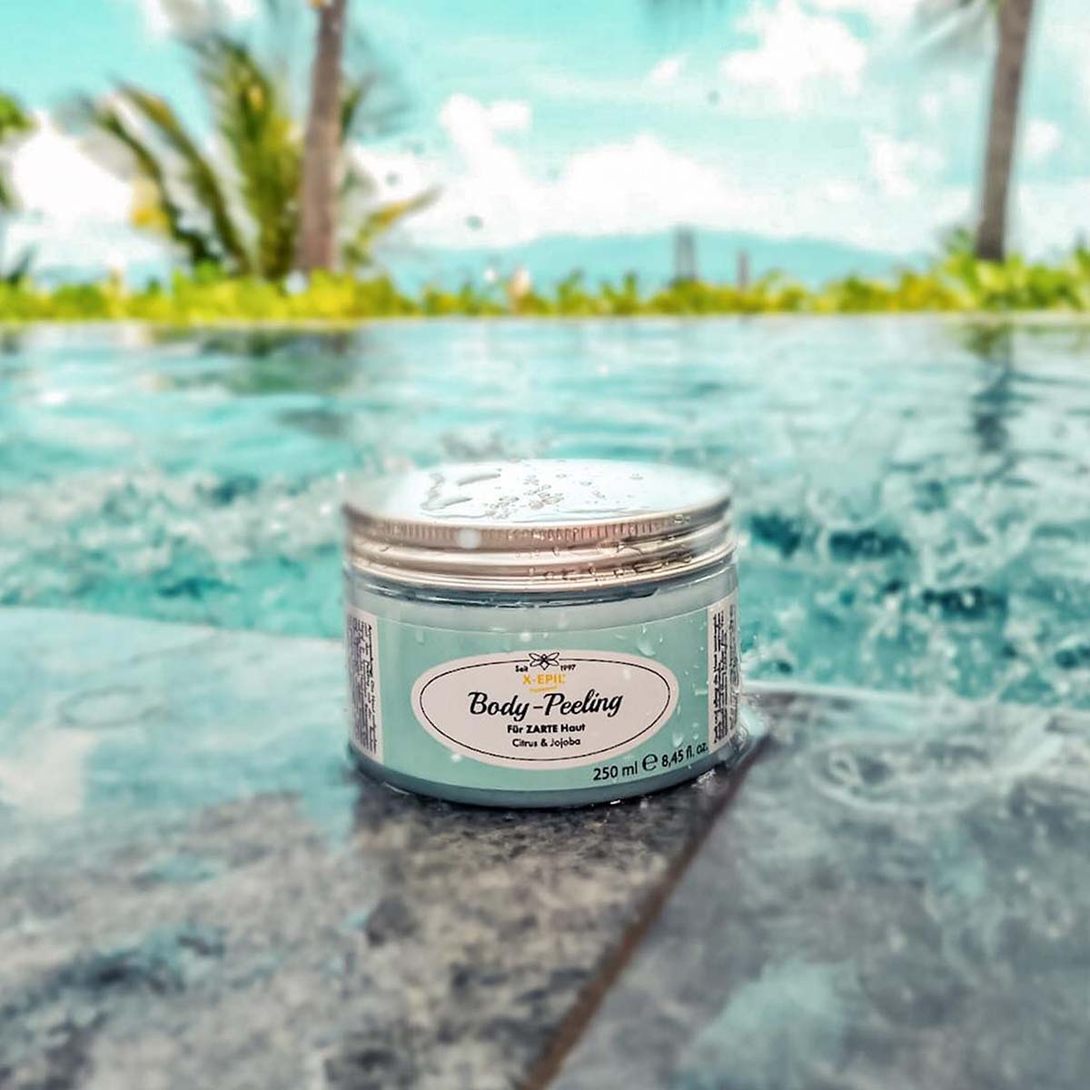 X-EPIL Body-Peeling Zitrus & Jojoba – 250ml, Tiegel an Poolrand mit Palmen