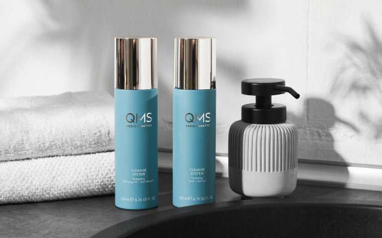 Badezimmeroberfläche neben einem Waschbecken mit zwei QMS Produkten aus der Cleanse System Serie: einem Cleansing Gel und einem Hydrating Toner. Daneben steht eine Seifenspenderflasche, im Hintergrund liegen zwei gefaltete weiße Handtücher.