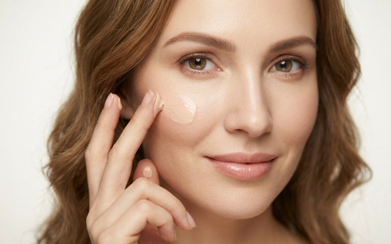 Nahaufnahme einer brünetten Frau mit langen Haaren und grünen Augen, die CC Cream mit einer Hand auf ihr Gesicht aufträgt. Eine Gesichtspartie ist mit der hautfarbenen CC Cream bedeckt, die sich dem natürlichen Hautton anpasst. Die Frau lächelt leicht.