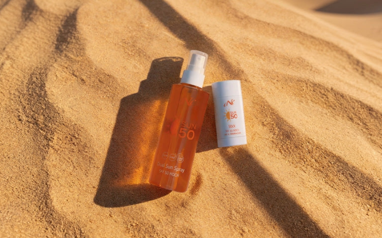 Zwei Sonnenschutzprodukte von CNC liegen auf gelbem Sand: eine Flasche Dual Sun Spray SPF 50 und daneben ein Sun Stick SPF 50.
