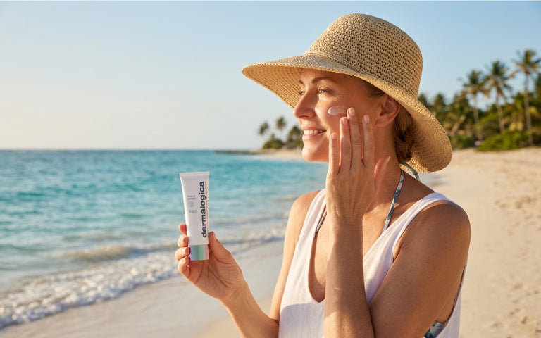 Frau im Profil steht an einem Sandstrand mit Sonnenhut und Badebekleidung, während sie Sonnencreme auf ihr Gesicht aufträgt. In der anderen Hand hält sie die Tube Prisma Protect SPF 30 von Dermalogica. Im Hintergrund sind das Meer und Palmen zu sehen.