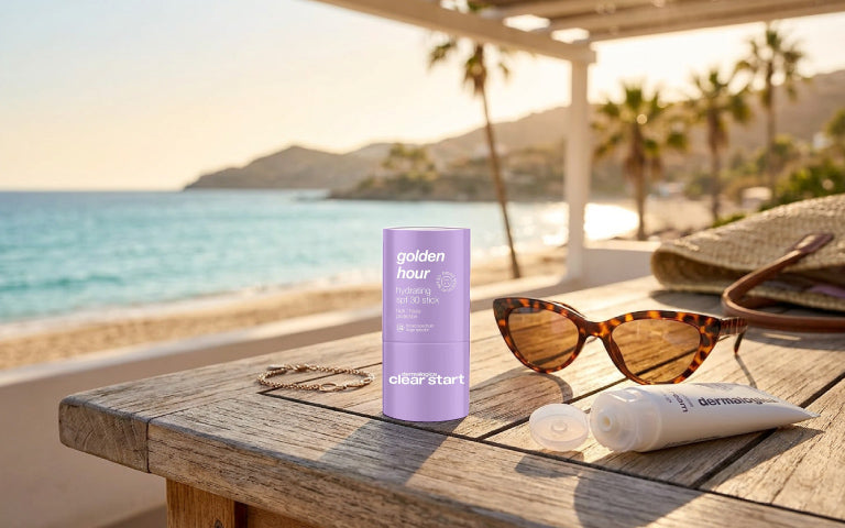 Auf einem Holztisch stehen Sonnenschutzprodukte von Dermalogica: der Golden Hour Hydrating SPF 30 Stick und eine geöffnete Tube Sonnencreme. Daneben liegen eine Sonnenbrille, ein goldener Armreif und ein Teil einer Strohtasche. Im Hintergrund sind ein Sandstrand, das Meer, Berge und Palmen zu sehen.