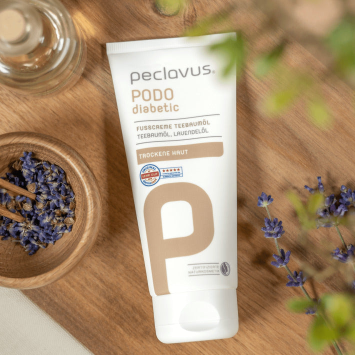 PECLAVUS Fußcreme Teebaumöl  – 100ml, Tube liegend auf Holz mit Lavendelblüten
