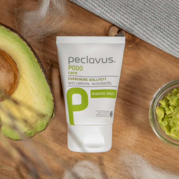 peclavus Fußcreme Wollfett  – 100ml, Tube auf Holz liegend mit augeschnittener Avocado