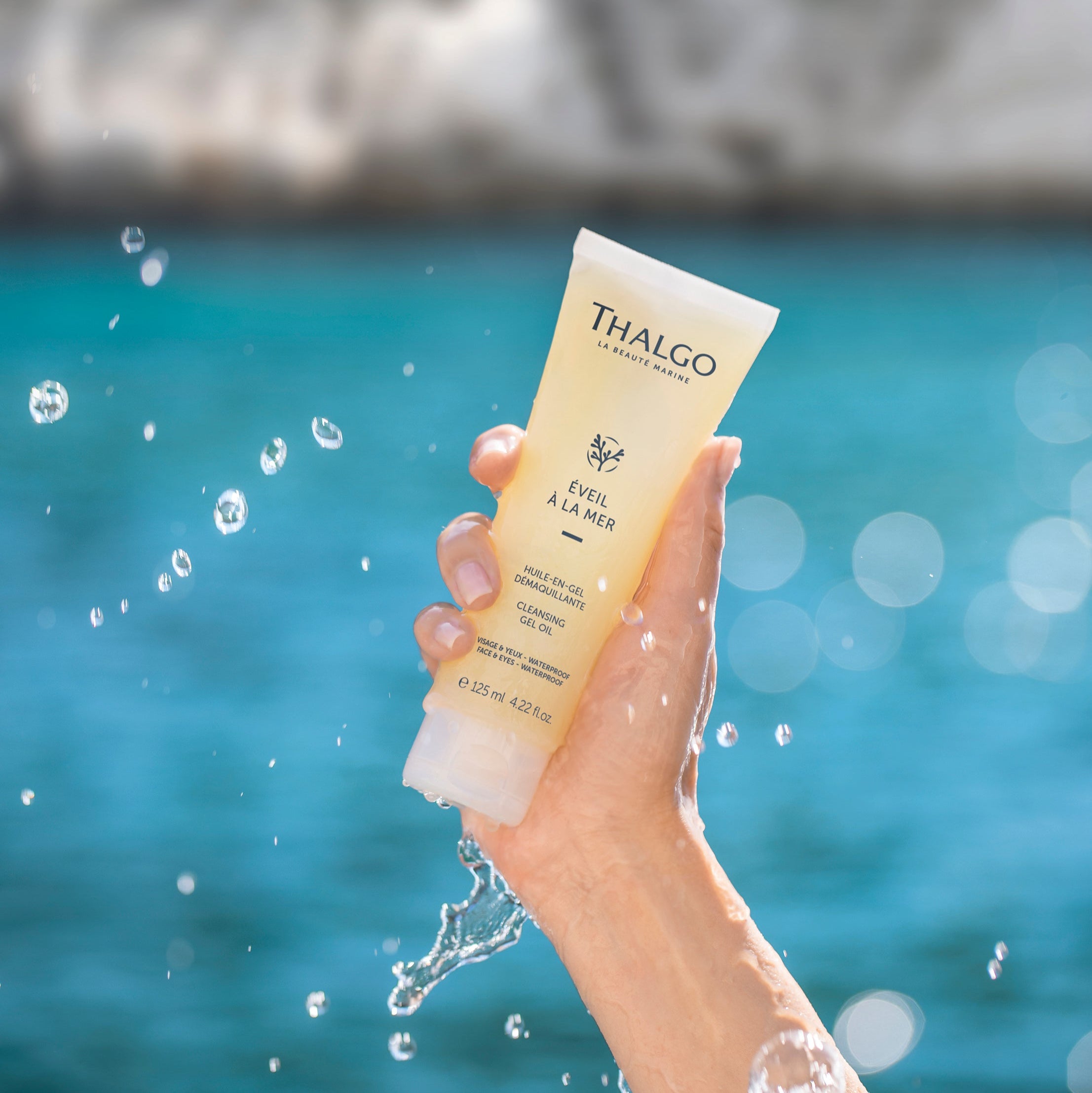 THALGO Éveil à la Mer - Reinigungsöl-Gel  – 125ml, Hand hält Tube mit Wassertropfen vor Hintergrund aus Wasser