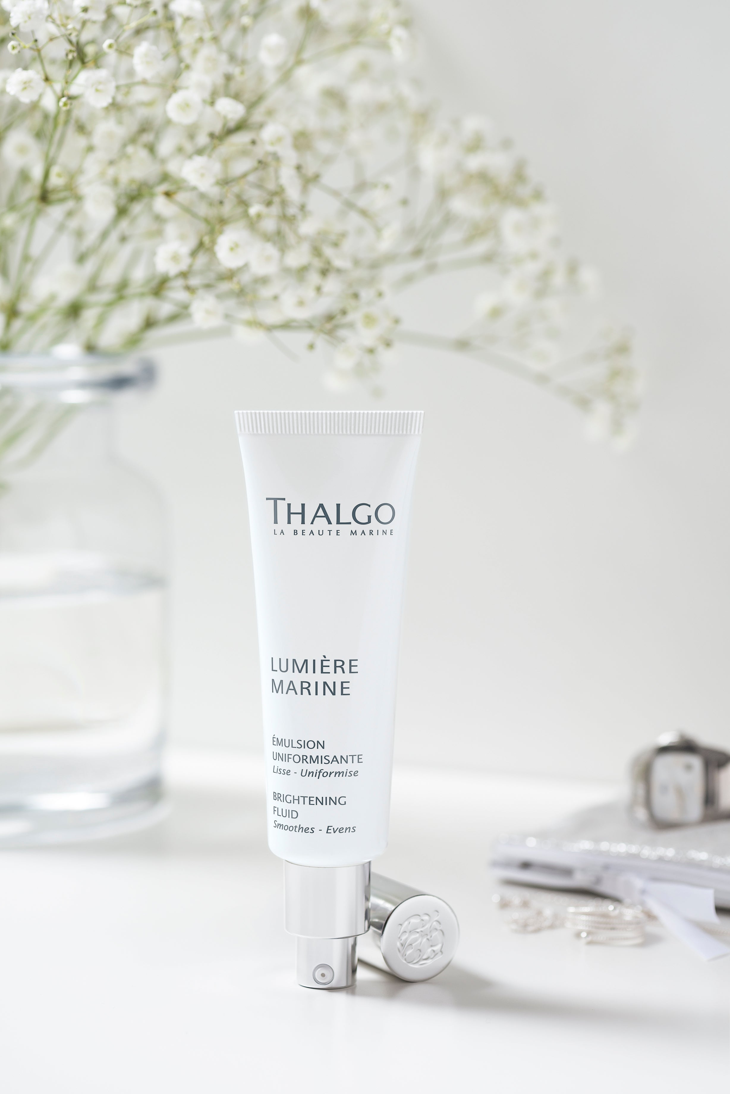 THALGO  Lumière Marine - Brightening-Fluid  – 50ml, Tube auf Tisch mit Schmuck und Blumenstrauß