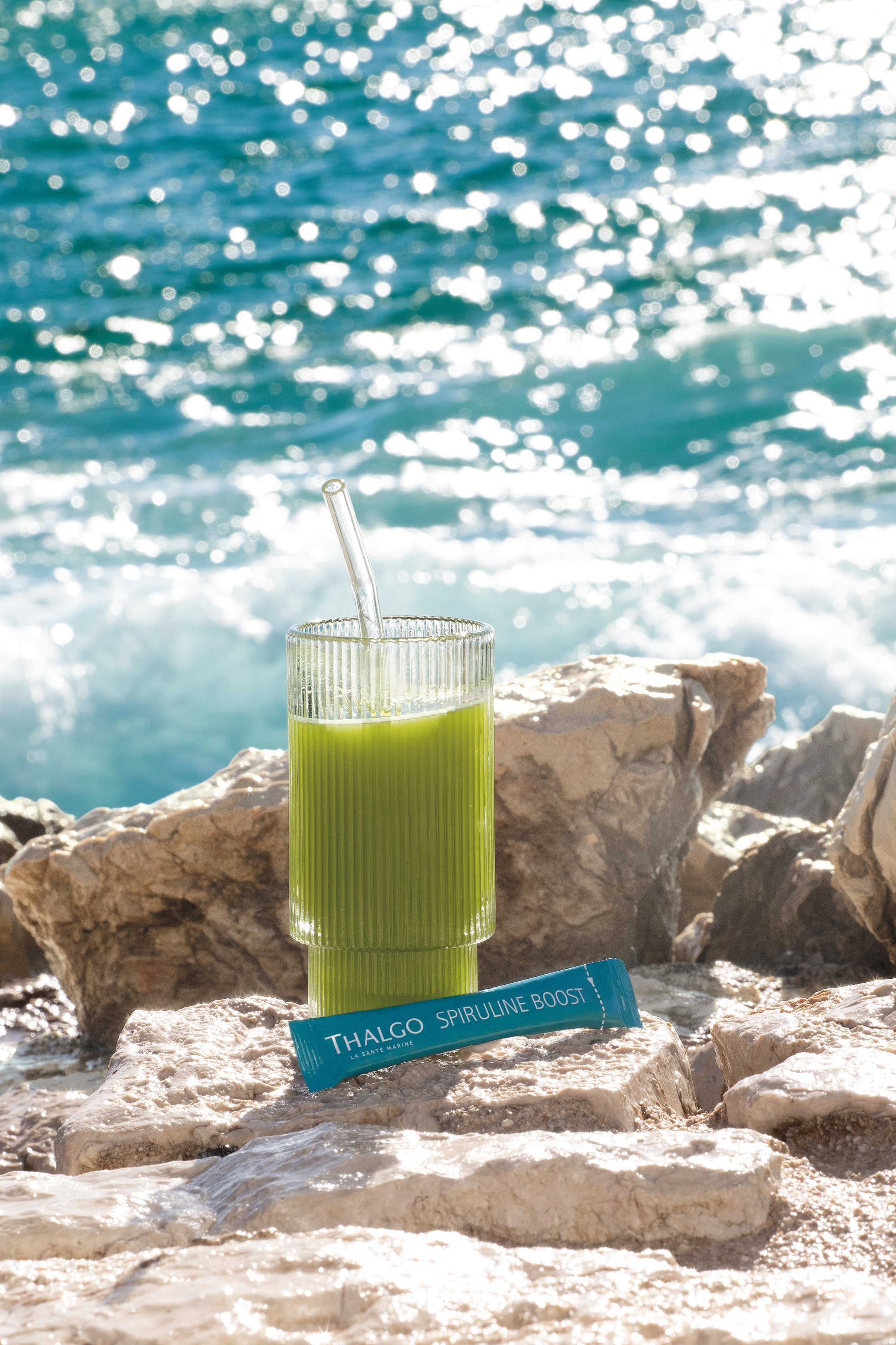 THALGO Spirulina Boost  – 5g, Einzelsachet und Spirulina-Shake im Glas mit Röhrchen auf Steinküste